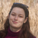 a dark-haired person wears a red turtleneck and has sunglasses on their head; they smile at the camera