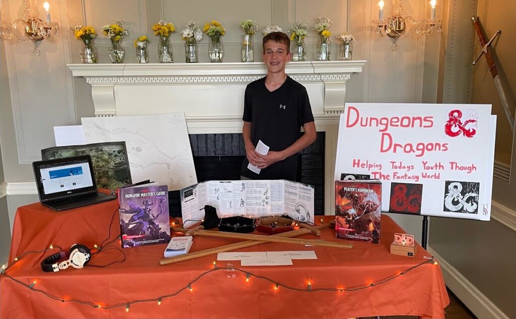 a youth stands before a table covered by a red tablecloth, displaying Dungeons & Dragons books, dice in a dice tray, and a poster that reads "Dungeons & Dragons: Helping Today's Youth Through The Fantasy World"
